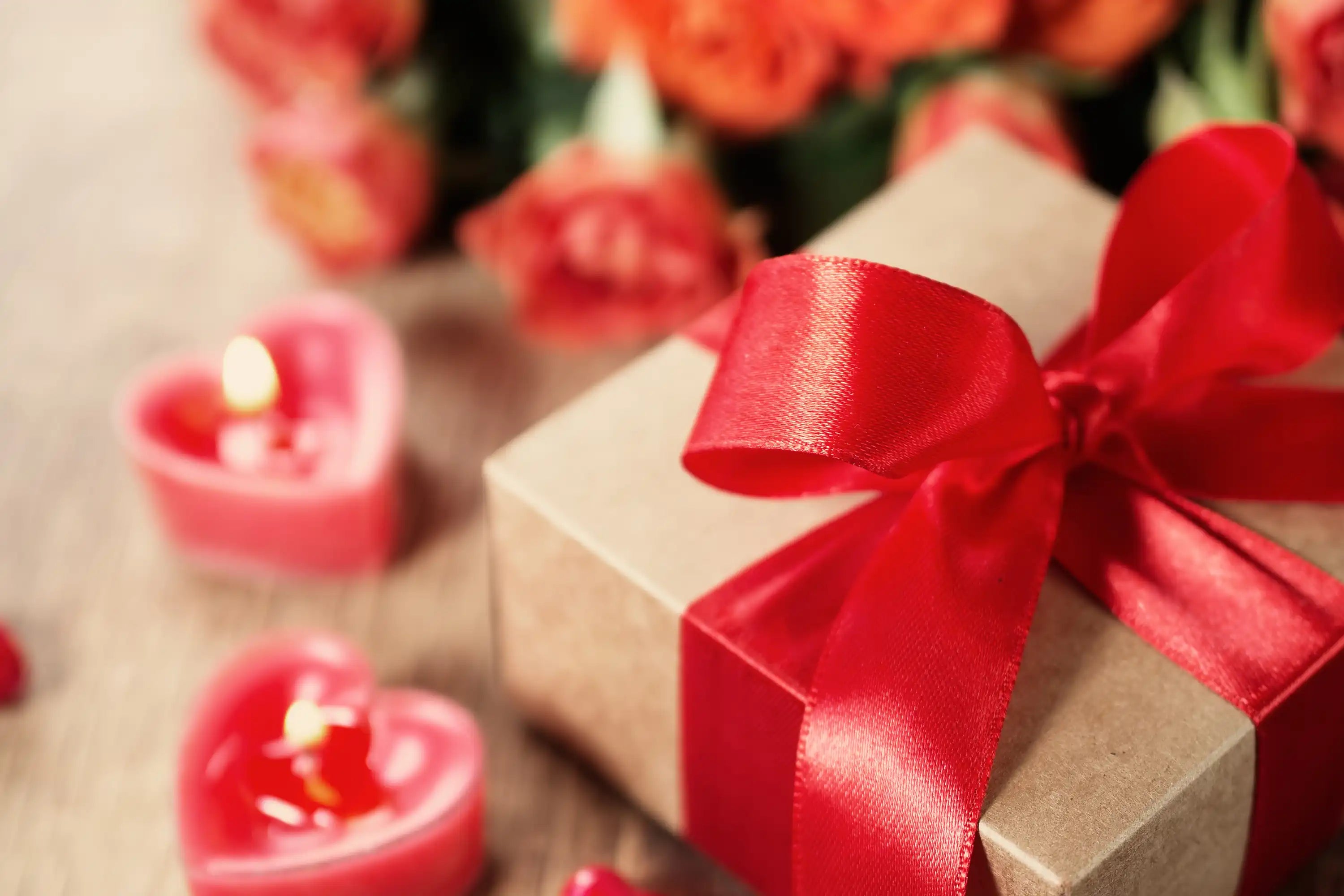 Geschenkkarton mit rotem Band, herzförmigen Kerzen und verschwommenen Blumen im Hintergrund auf einer Holzoberfläche.