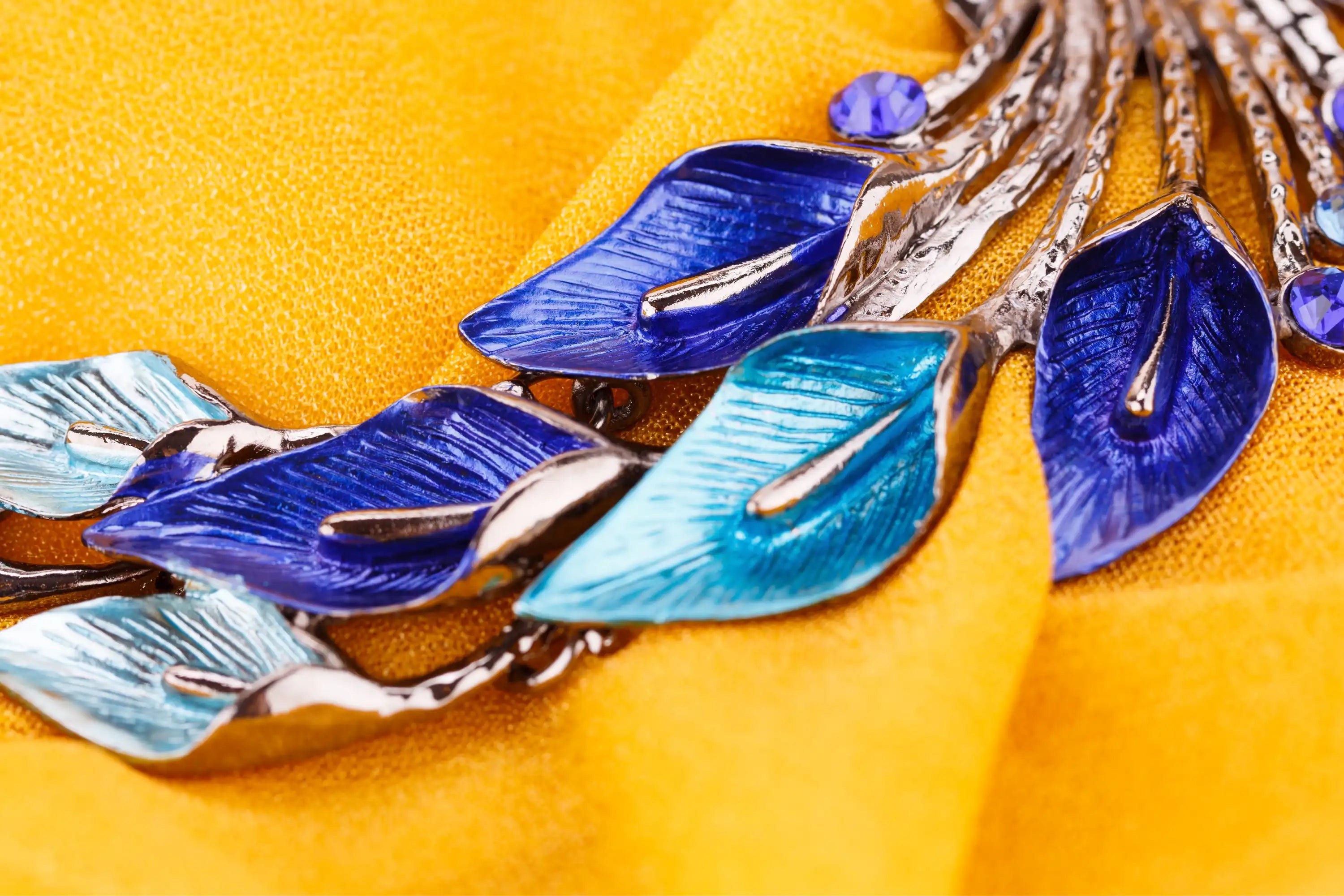 Nahaufnahme einer blauen und türkisfarbenen blattförmigen Brosche auf leuchtend gelbem Stoff.