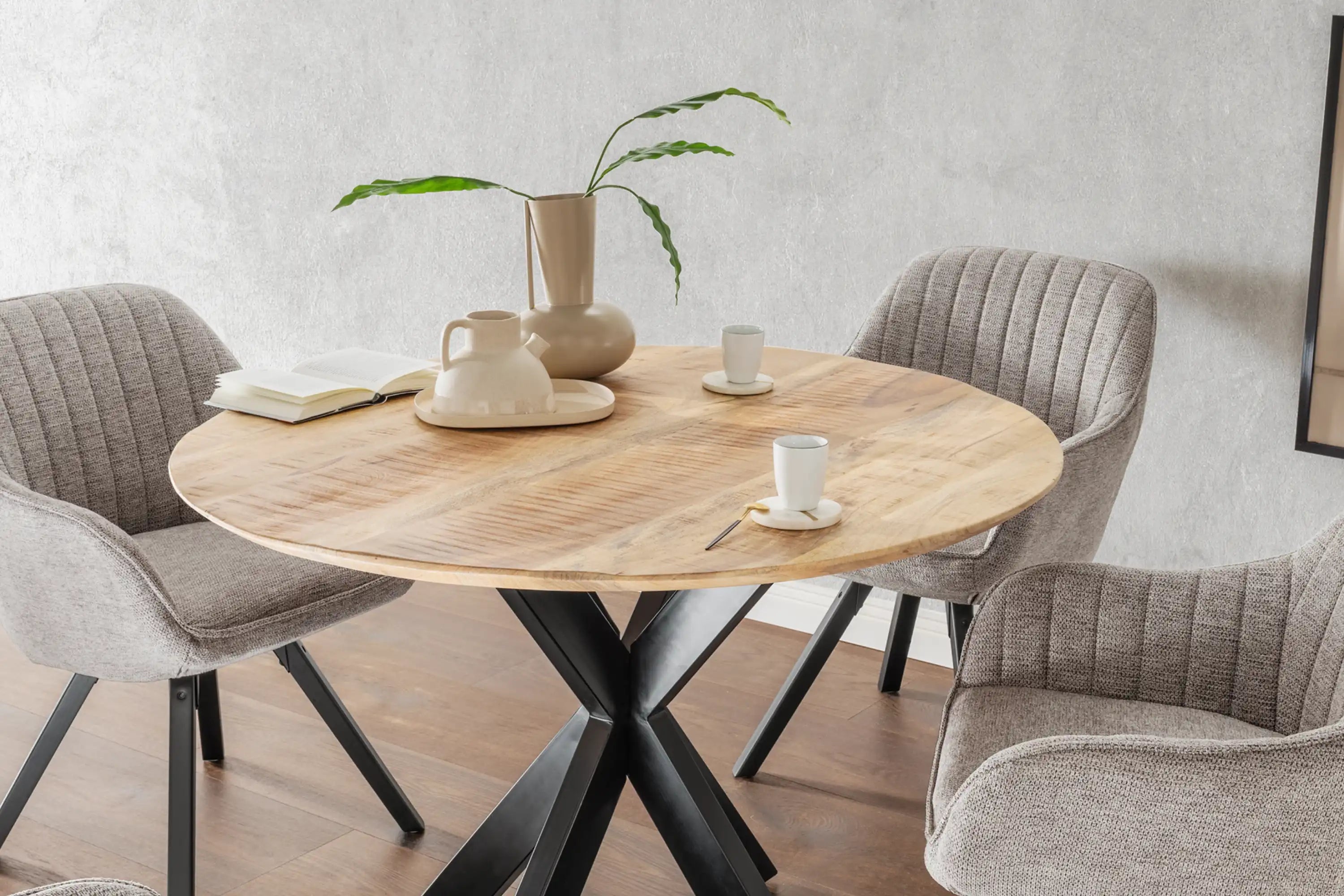 Runder Holztisch mit beigen Stühlen, einer Vase mit grünen Blättern, Tassen und einem Buch darauf.