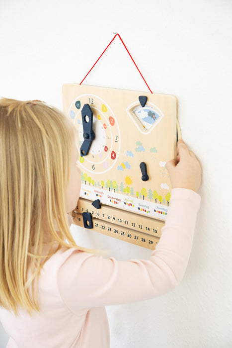 Ein junges Mädchen interagiert mit einer an der Wand montierten Lernuhr und Kalendertafel von Holzspielzeug und verbessert so ihr Zeitverständnis und entwickelt gleichzeitig ihre Feinmotorik.