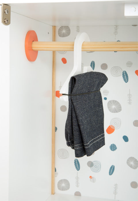 Eine dunkle Jeans hängt an einem weißen Haken in einem Schrank mit gemusterter Tapete und ähnelt einem Puppenmöbelstück in einem Kleiderschrank aus Holz.