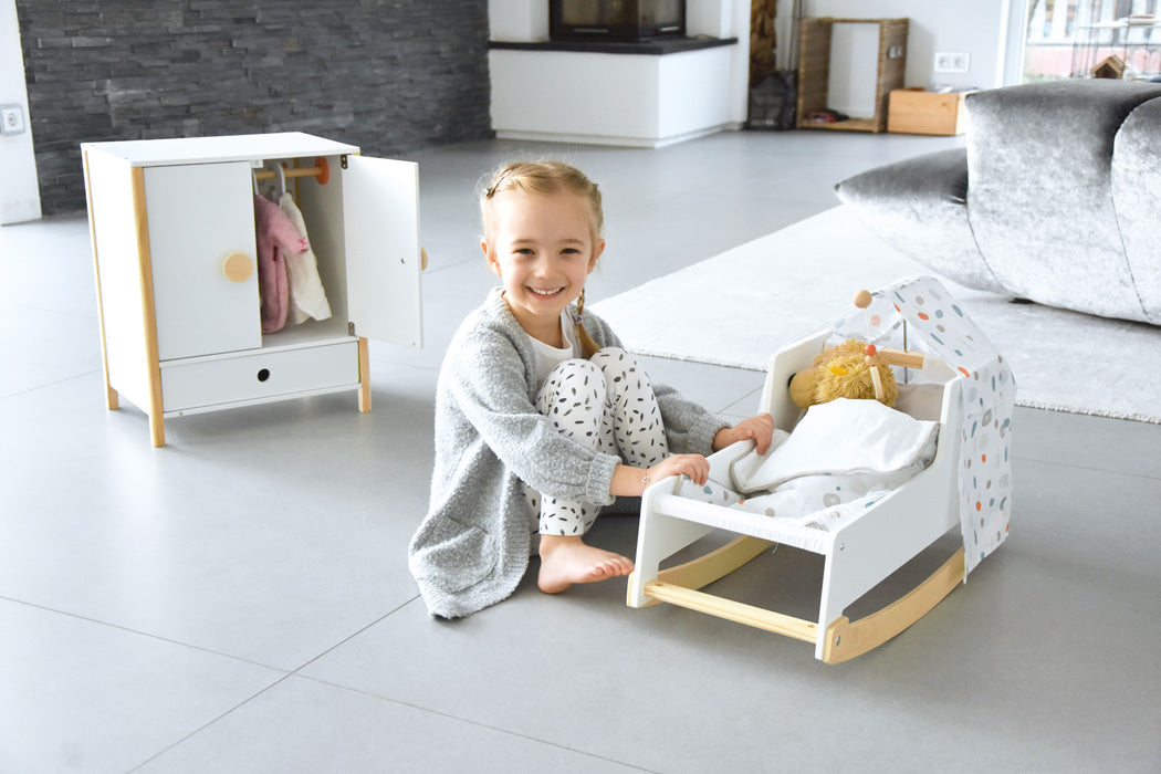 Lächelndes Mädchen sitzt auf dem Boden neben einer Puppenwiege und einem kleinen Kleiderschrank in einem modernen Wohnzimmer mit skandinavischem Design.