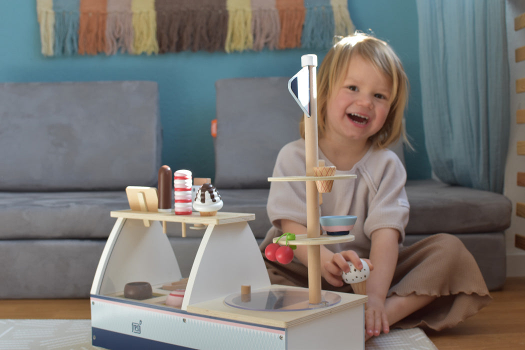 Lächelndes Kind, das in einem gemütlichen Wohnzimmer mit einem hölzernen Eis-Spielset und einem magnetischen Eisportionierer auf dem Boden spielt.