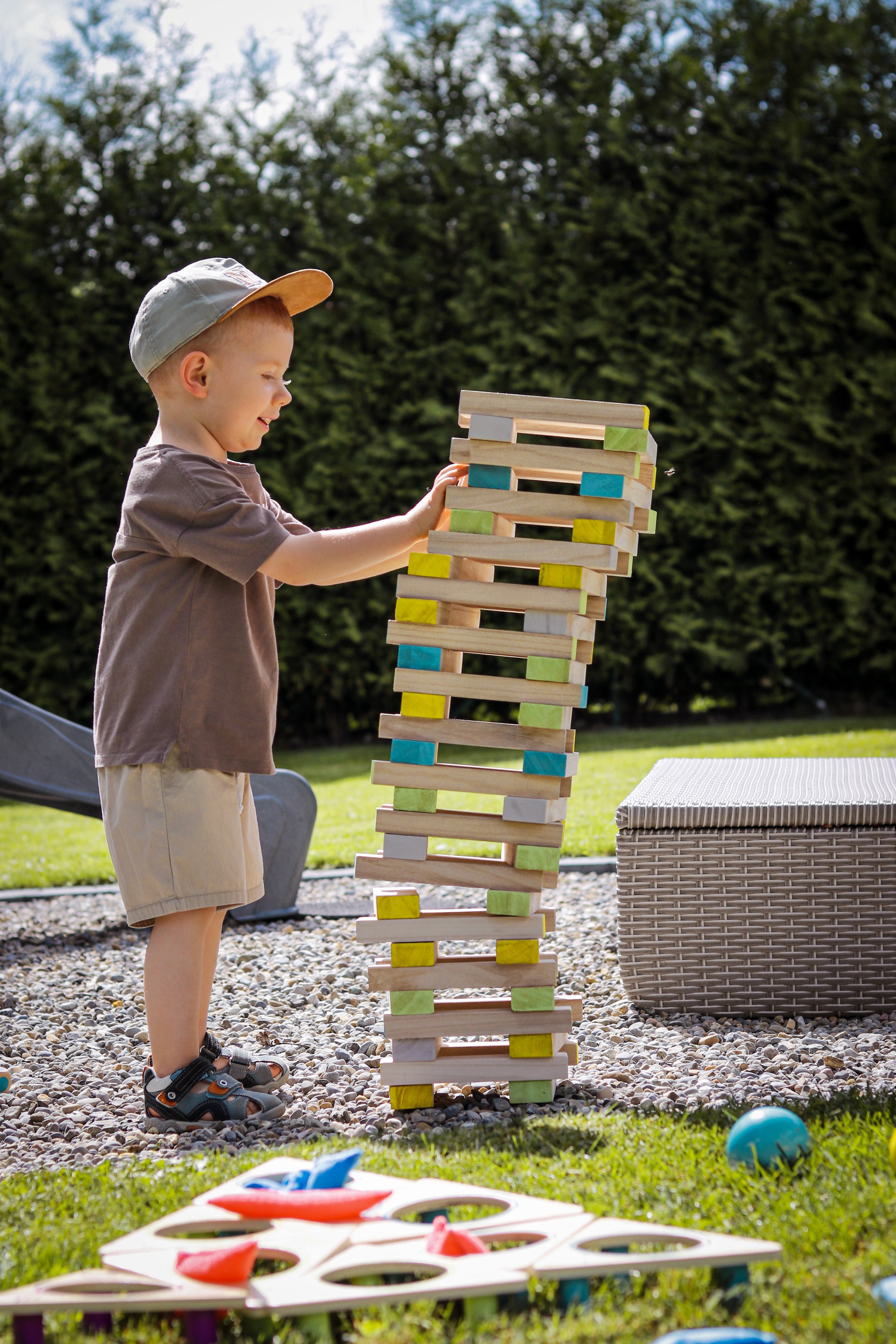 Ein kleiner Junge stapelt im Freien sorgfältig FSC® 100 %-zertifizierte Holzklötze und baut einen hohen, bunten Wackelturm-Spielturm, der kurz vor dem Umfallen steht.