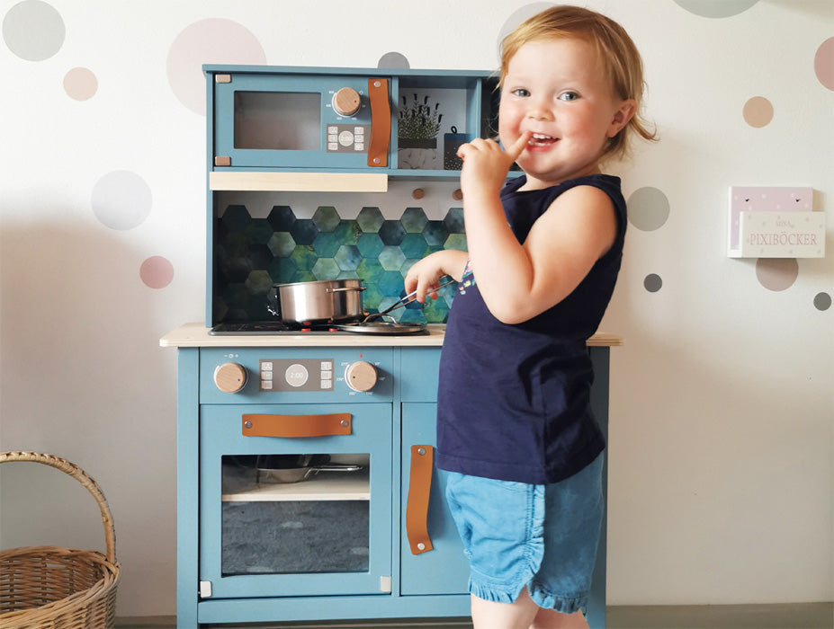 Ein lächelndes Kleinkind steht an einer rauchblauen Spielküche, hält einen Löffel und tut so, als würde es kochen, wodurch die Spielzeit in einen bezaubernden Designobjekt-Moment verwandelt wird.