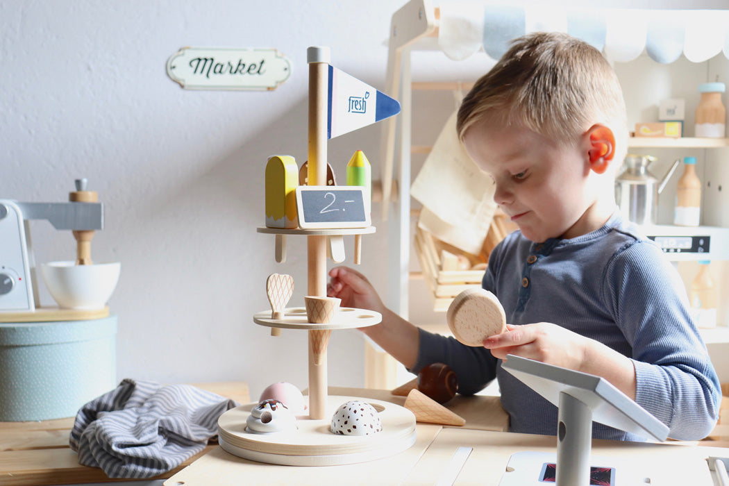 Kleiner Junge spielt mit einem Eisständer aus Holz und einem Eiskugelportionierer auf einem vorgetäuschten Markt oder in einer Küche und genießt das kreative Rollenspiel Eis.