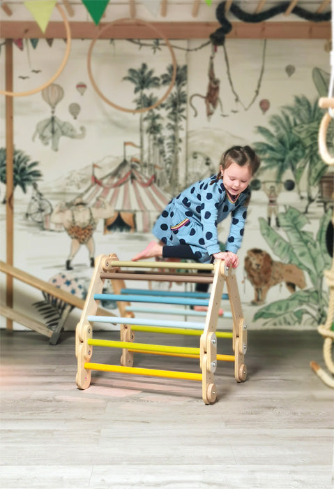 Ein junges Mädchen in einem blauen Outfit klettert in einem farbenfrohen Indoor-Spielzimmer auf ein hölzernes Kletterdreieck und genießt Kreativität und Abenteuer.