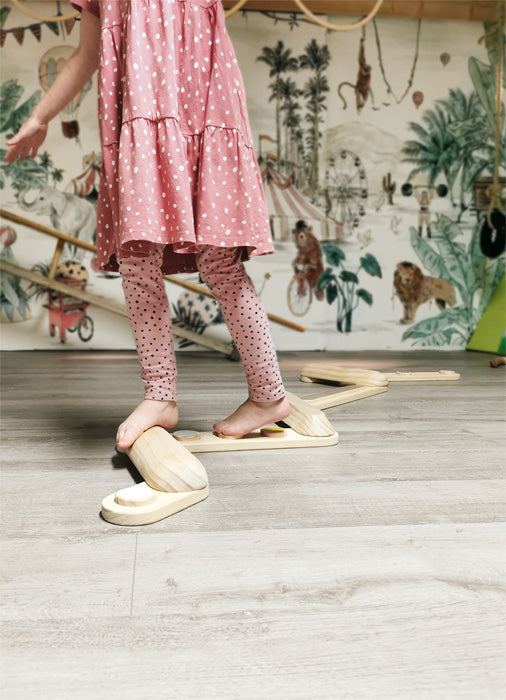 Kind im rosa Kleid und gepunkteten Leggings balanciert auf Balancierpfaden im Innenbereich mit Tiertapete und entwickelt spielerisch seinen Gleichgewichtssinn.