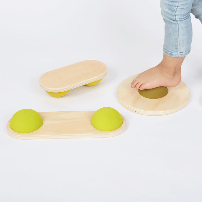 Der nackte Fuß eines Kindes berührt ein hölzernes Balancespielzeug (Balanciersteine), wodurch der Gleichgewichtssinn und die Koordination zwischen ähnlichen Spielzeugen auf einer weißen Oberfläche verbessert werden.