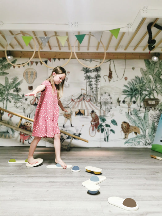 Ein junges Mädchen in einem rosa Kleid balanciert auf Balanciersteinen in einem Spielzimmer mit einem Wandgemälde zum Thema Dschungel und verbessert so spielerisch ihre Koordination und ihren Gleichgewichtssinn.
