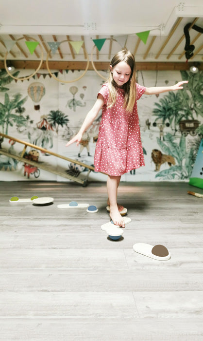 Ein junges Mädchen in einem rosa gepunkteten Kleid balanciert auf Balanciersteinen in einem Spielzimmer mit einem Dschungel-Wandbild und verbessert so ihre Koordination und ihren Gleichgewichtssinn.