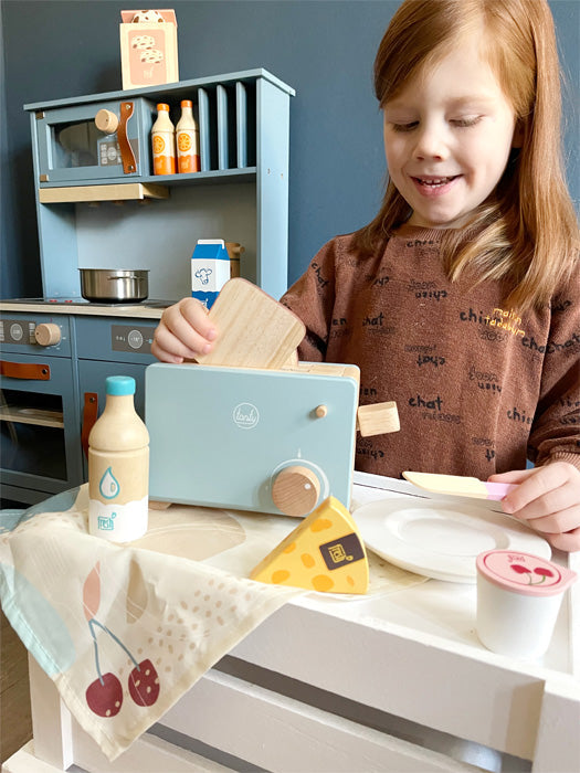 Ein lächelndes Kind spielt mit einem Toaster und hölzernem Lebensmittelzubehör in einer entzückenden Kinderküche.