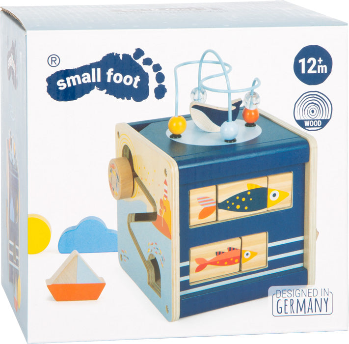 Blaues Aktivitätswürfel-Spielzeug aus Holz mit Fischmotiven, Perlen und einem „12+ Monate“-Etikett auf der Verpackung – dieser Motorikwürfel bietet spannendes Spielen für die Kleinen.