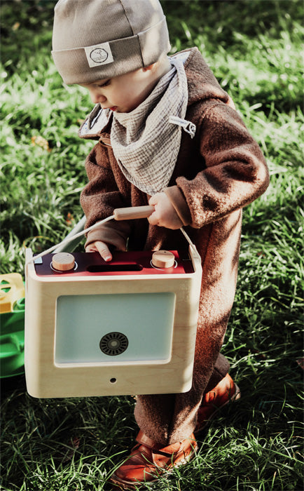 Ein kleines Kind in braunem Outfit hält ein hölzernes Spielzeugradio in der Hand, während es auf grünem Gras neben einem Campingkoffer für die multifunktionale Spielküche steht.