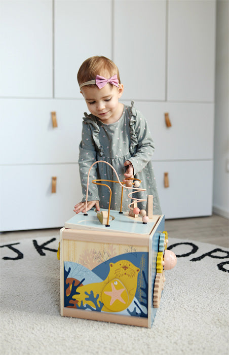 Ein Kleinkind in einem grünen Kleid spielt mit einem Motorikwürfel auf einem Teppichboden, erkundet dieses Holzspielzeug und entwickelt seine Feinmotorik-Fähigkeiten.