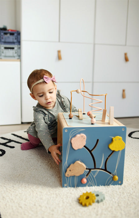 Ein Kleinkind mit einem rosa Stirnband mit Schleife entwickelt Feinmotorik, während es mit einem Motorikwürfel von Holzspielzeug auf einem Teppichboden spielt.
