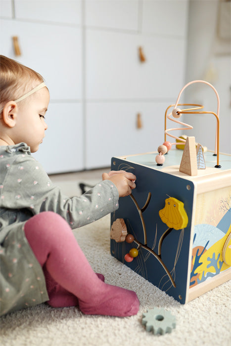 Ein Kleinkind spielt mit einem Motorikwürfel aus Holzspielzeug auf einem Teppichboden in einem hellen Raum und erkundet und entwickelt dabei seine Feinmotorik.