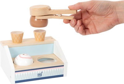 Hand hält eine Holzzange über einem Eiscreme-Spielset mit Waffeln und Kugeln auf einem kleinen Präsentationsständer – perfekt für die Kinderküche und fantasievolles Spielen mit dem magnetischen Eisportionierer.