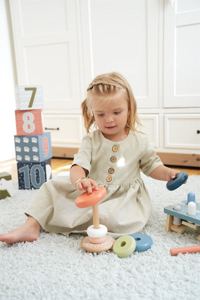 Ein Kleinkind spielt mit einem Stapelturm aus FSC-zertifiziertem Holz auf einem Teppich, mit nummerierten Blöcken im Hintergrund, was Spaß und gesundes Motorikspielzeugspiel fördert.