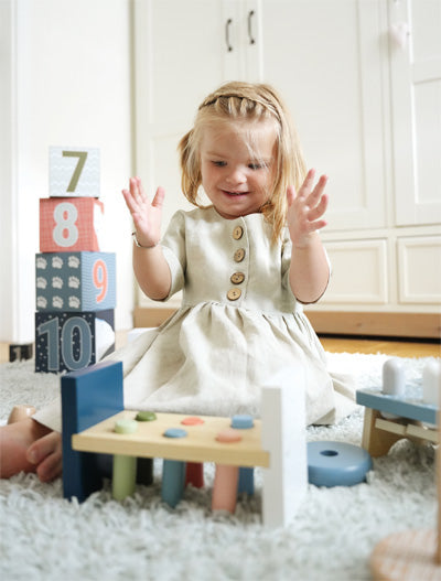 Junges Mädchen in hellem Kleid spielt auf dem Boden mit Stapelklötzen und einer Hammer- und Klopfbank und stärkt so ihre motorischen Fähigkeiten.