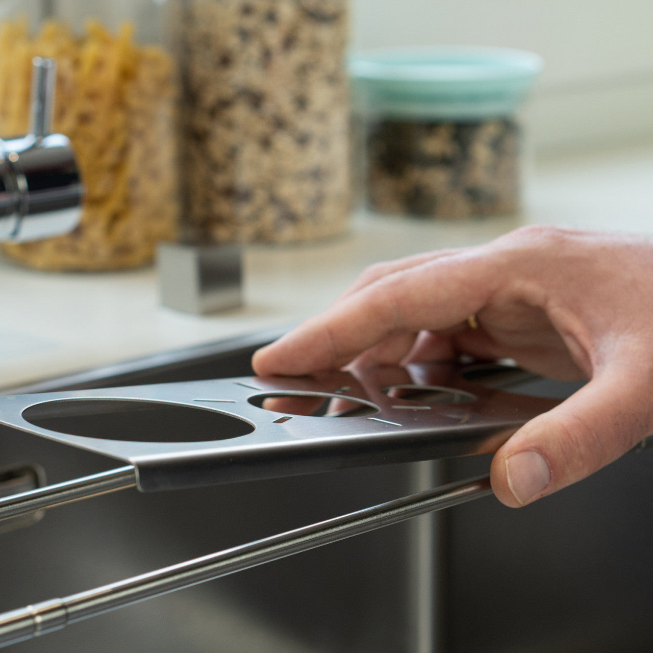 Eine Hand hält ein silbernes Nudelmessgerät aus Metall mit Edelstahleinsatz über einer Küchenspüle.