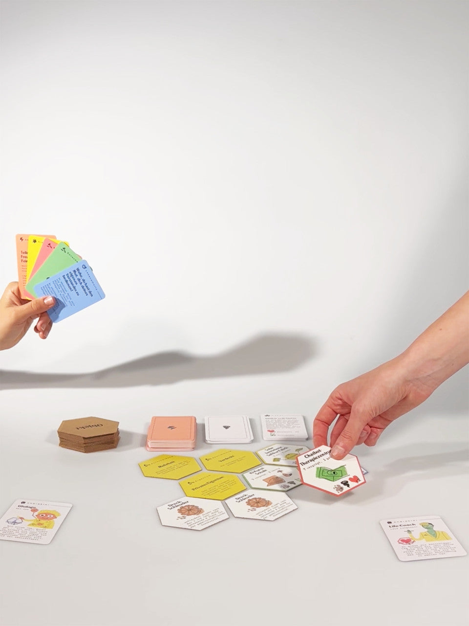 Zwei Hände spielen als Weltraumreisende ein Gesellschaftsspiel mit bunten Karten und sechseckigen Spielsteinen auf einer weißen Oberfläche und erwecken das Gefühl, einen verlassenen Planeten zu erkunden.