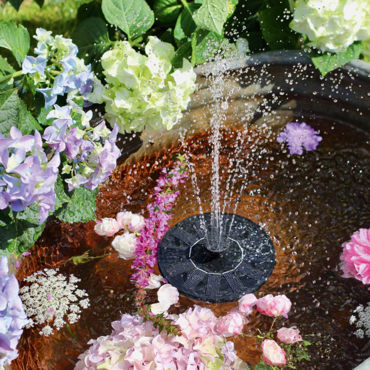 Ein kleiner Solar-Brunnen sprüht Wasser in ein Vogelbad im Garten, umgeben von bunten Blumen und grünen Blättern, und fügt Ihrer Gartendeko ein bezauberndes Wasserspiel hinzu.