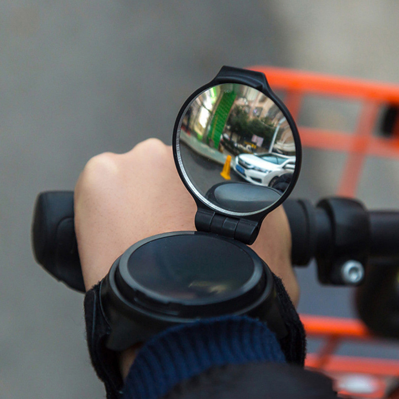 Eine Hand am Fahrradlenker trägt einen Zusatzspiegel am Handgelenk, der die Sicherheit im Straßenverkehr erhöht, indem er eine Auto- und Straßenszene reflektiert.