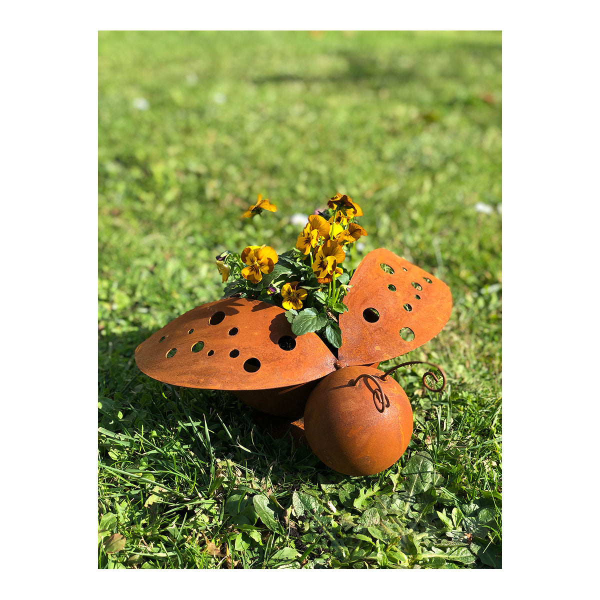 Ein metallener Marienkäfer-Gartenkübel in Edelrost-Optik, gefüllt mit gelben Blumen, steht im Sonnenlicht auf grünem Gras.