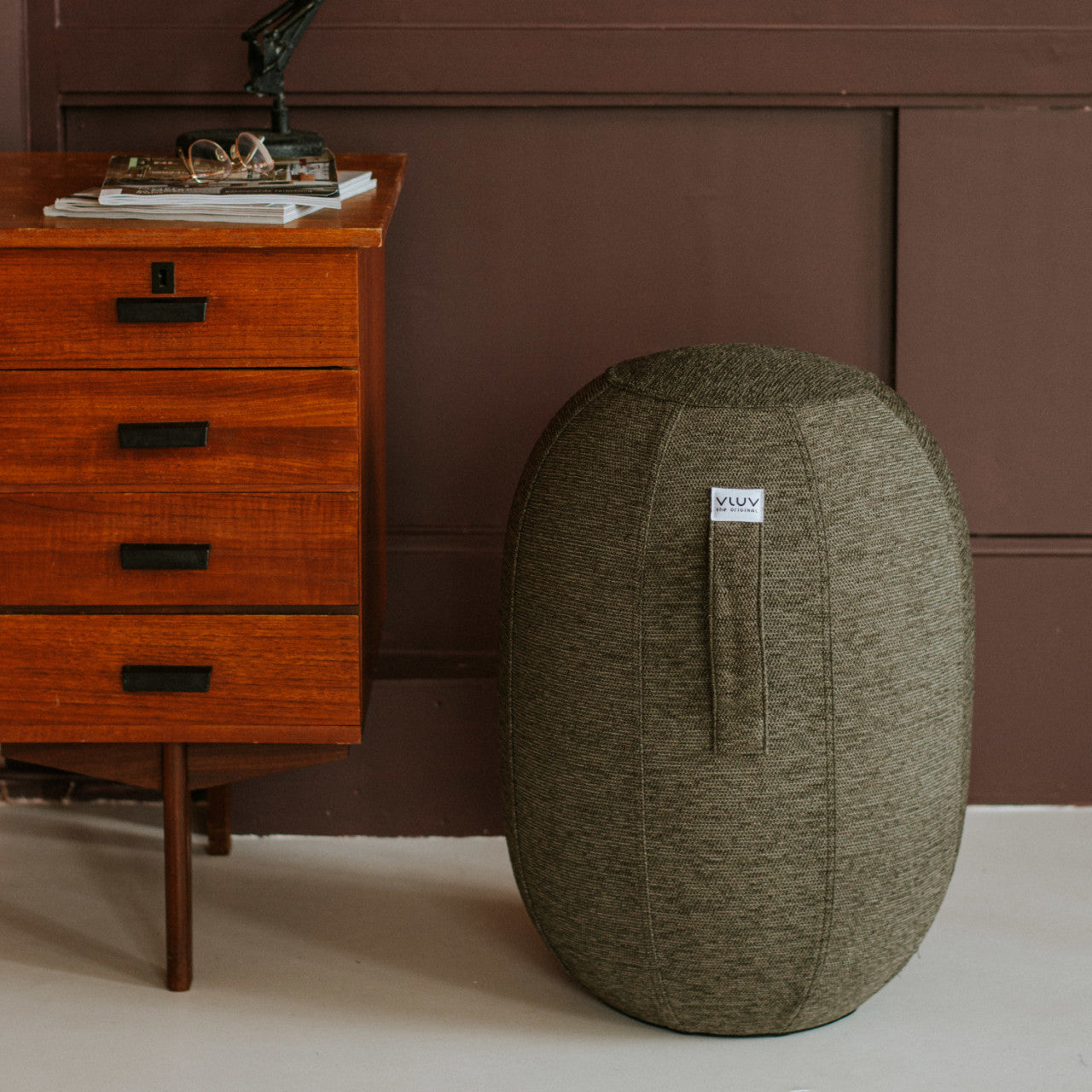 Grauer Balancehocker KAPSUL STOV neben einer Holzkommode mit Büchern und Lampe.