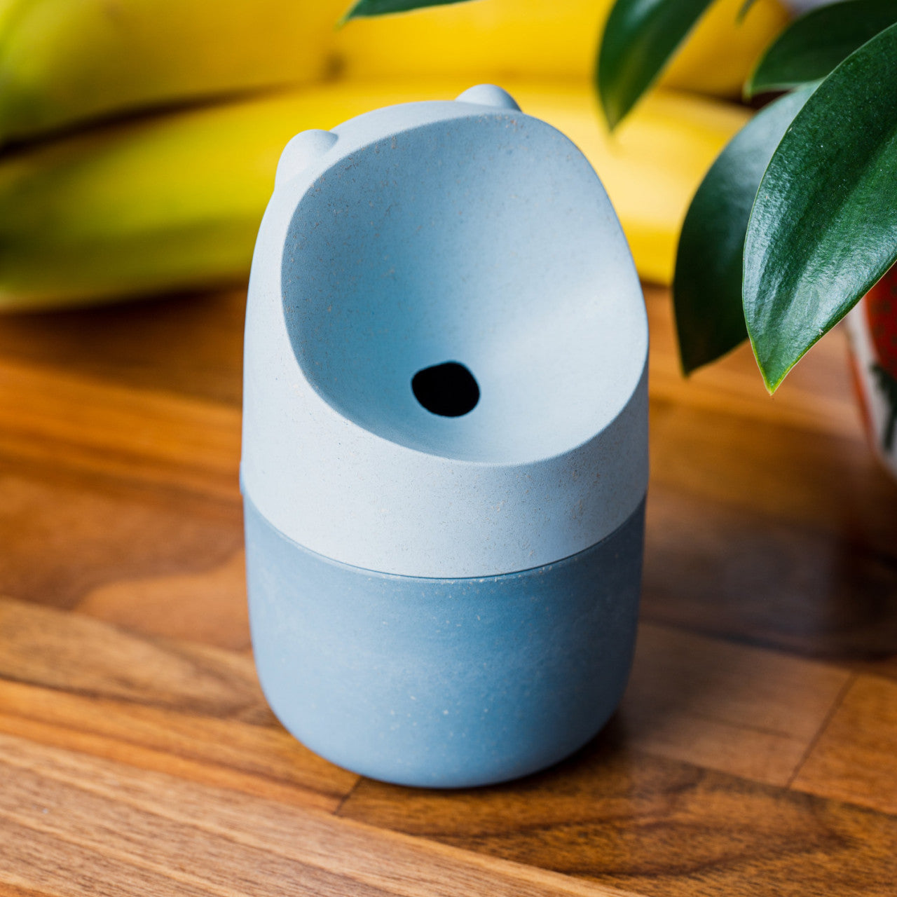 Eine kleine blaue Tasse mit Bärenohren aus umweltfreundlichem Holz-Bioverbundstoff steht auf einem Holztisch neben Bananen und grünen Blättern.
