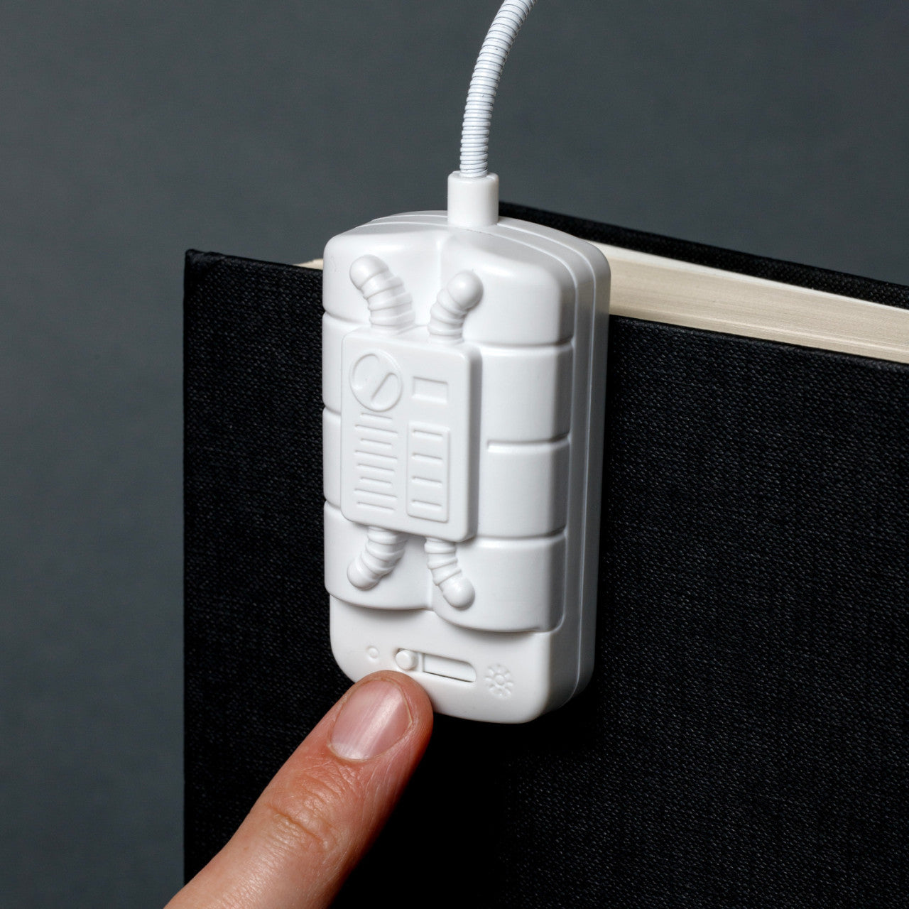 Ein Finger drückt auf eine weiße Leselampe, die am Einband eines schwarzen Buches vor einem grauen Hintergrund befestigt ist, und ist bereit, ihren fokussierten LED-Lichtstrahl zum Lesen abzugeben.