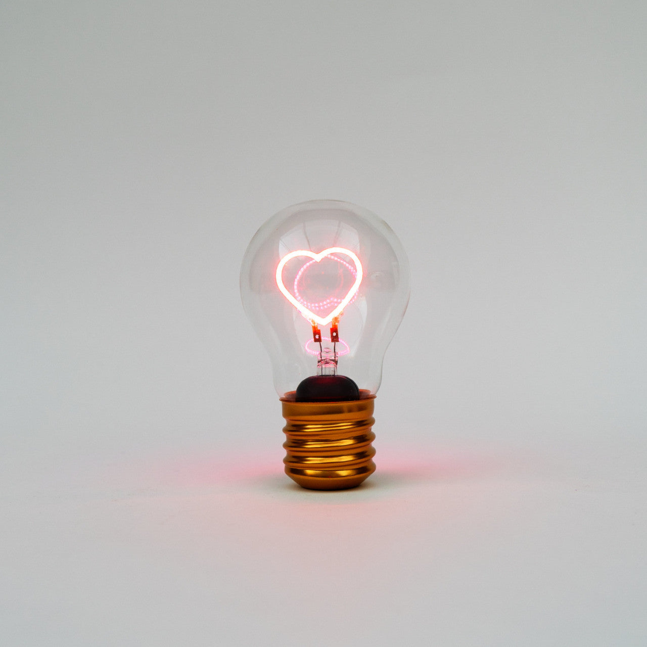 Klare Edison-Glühbirne mit leuchtendem herzförmigen Glühfaden vor einem schlichten weißen Hintergrund.