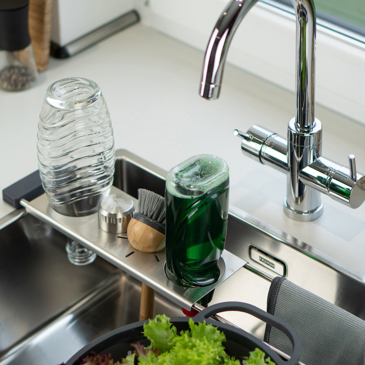 Glaswaren, eine Bürste und ein grüner Seifenspender auf einem Abtropfstation-Spülbeckenregal über einer Küchenspüle mit fließendem Wasser.