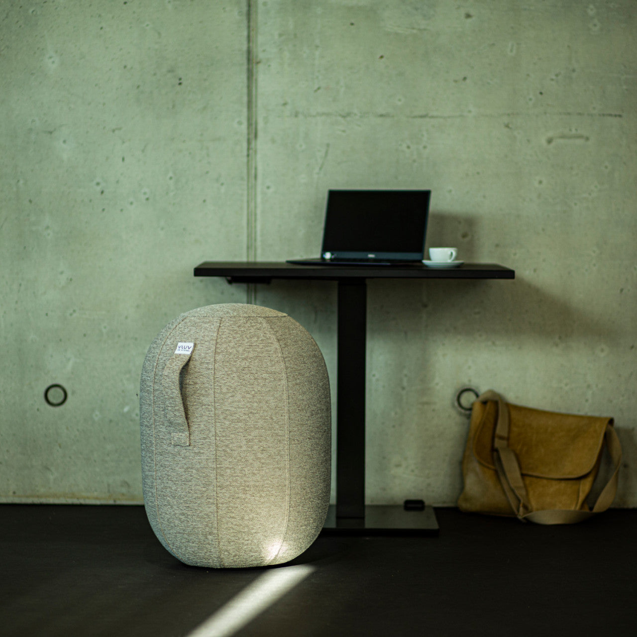 Ein Laptop und eine Tasse auf einem schwarzen Tisch, dazu eine beige Tasche und ein luftgefüllter Sitzhocker sorgen für ergonomisches Sitzen in einem minimalistischen Raum.