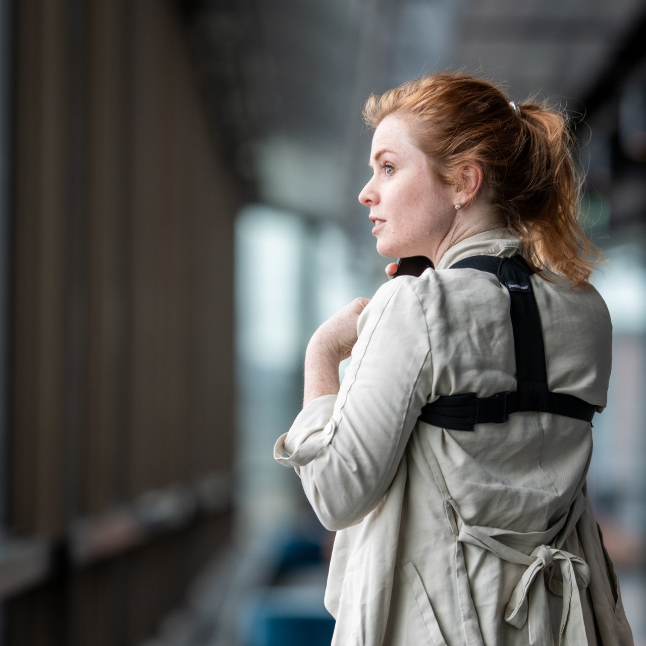 Frau mit roten Haaren, die eine Haltungsstütze trägt und aus einem großen Fenster im Innenbereich schaut.