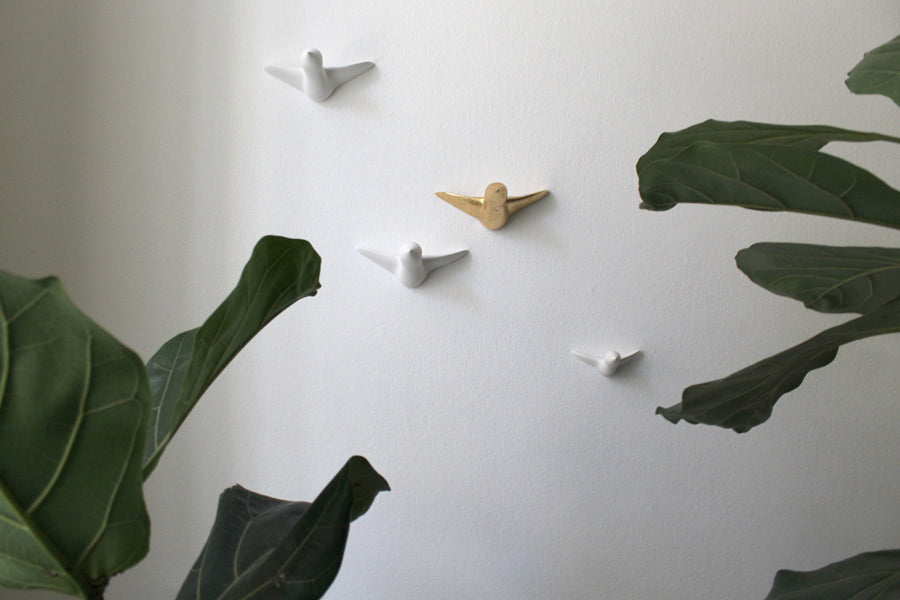 Vier minimalistische Wandskulpturen – kleine Vogelschwarme in Weiß und Gold – dienen gleichzeitig als funktionale Wandhaken an einer weißen Wand, eingerahmt von großen grünen Pflanzenblättern, die die abstrakte Komposition verstärken.