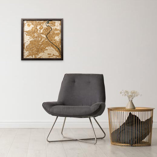 Ein grauer Sessel und ein runder Tisch mit einer Vase stehen unter einer gerahmten 3D-Holzwandkarte Stuttgart und machen die weiße Wand zu einem stilvollen Designobjekt.