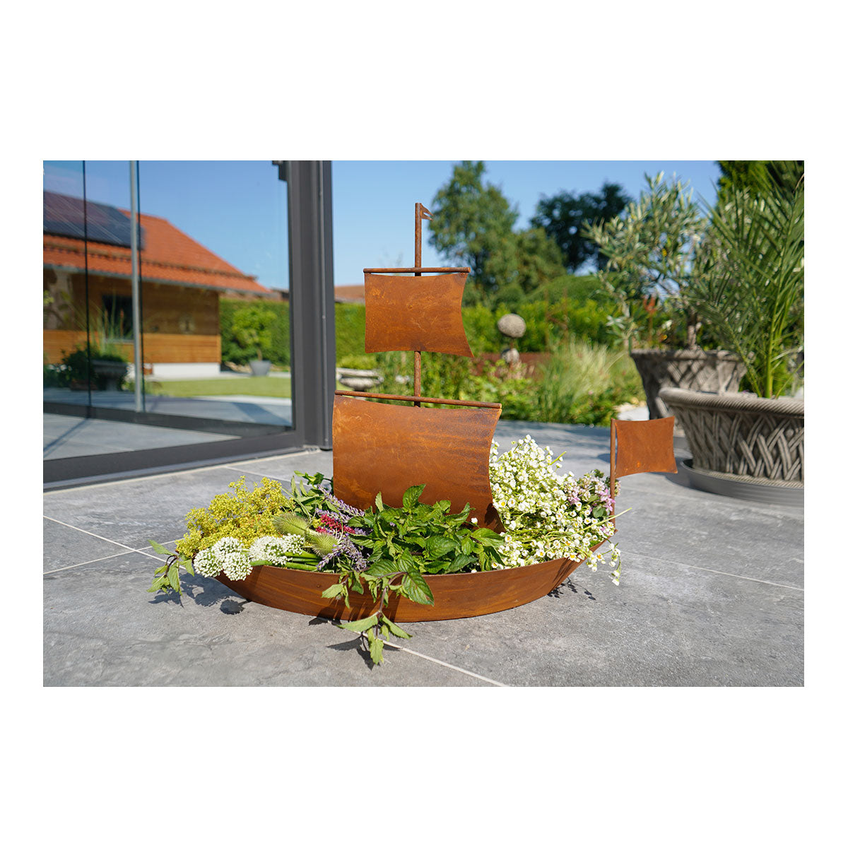 Schiffsförmiger Blumentopf aus Metall in Edelrost-Optik, gefüllt mit verschiedenen Blumen und Grünpflanzen, auf einer Terrassenplatte im Freien platziert – perfekt, um Ihrem Garten einen Hauch von Bayernwald-Charme zu verleihen.