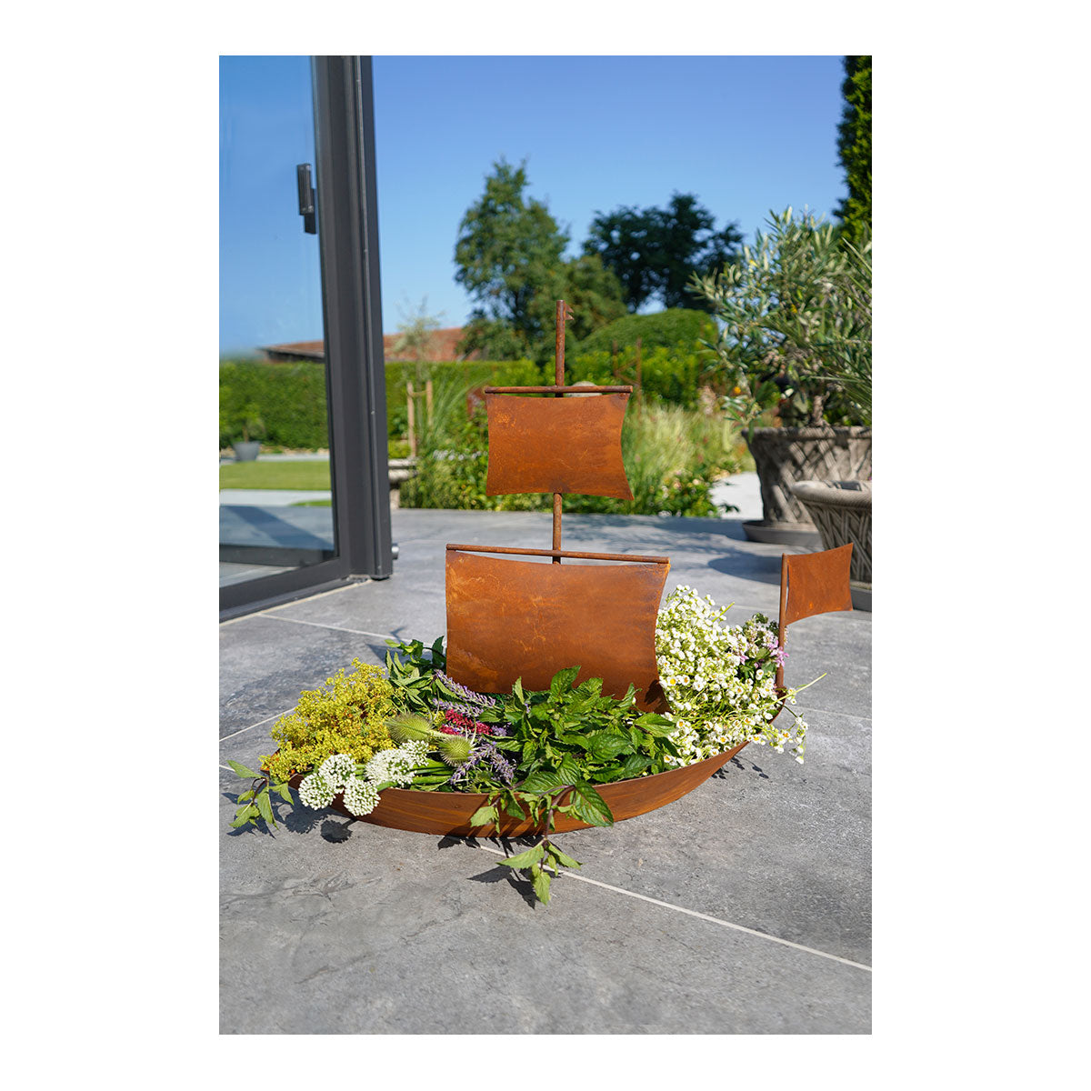 Dekoratives Metallobjekt in Edelrost-Optik: Dieses Metallschiff auf der Terrasse ist mit grünen Blättern und weißen Blumen gefüllt und steht vor einem üppigen Garten, der perfekt dazu geeignet ist, den Charme des Bayerischen Waldes zu verbreiten.