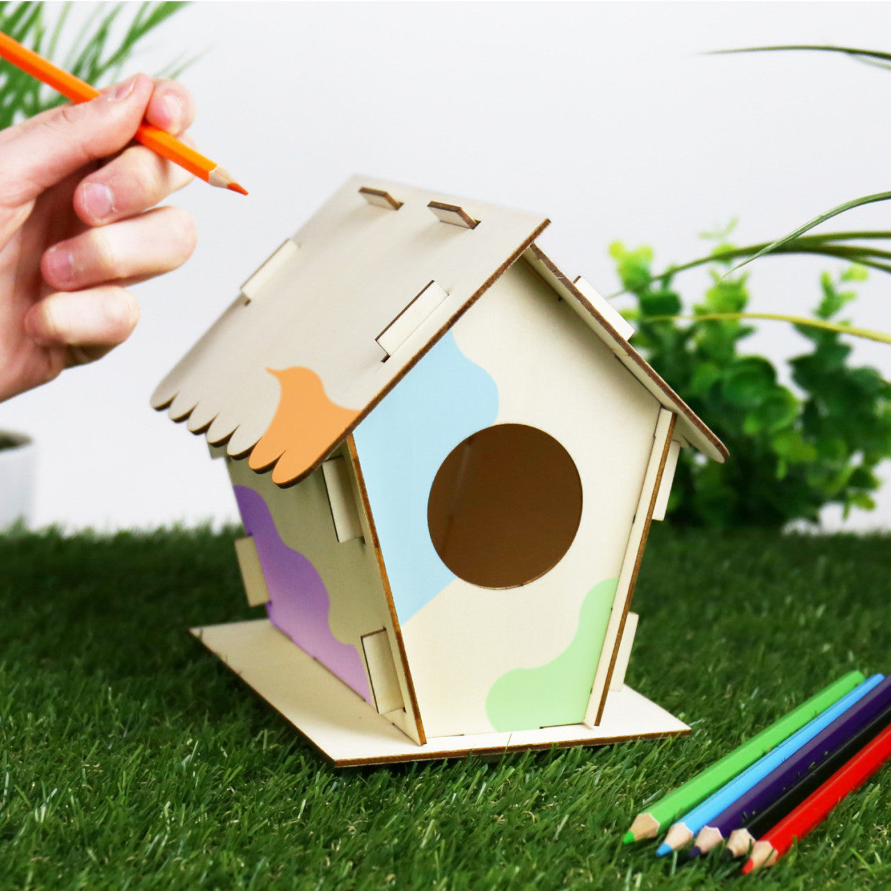 Eine Hand bemalt mit Pastellstiften ein hölzernes Vogelhaus „Vogelhotel“ auf Kunstrasen, im Hintergrund Pflanzen.