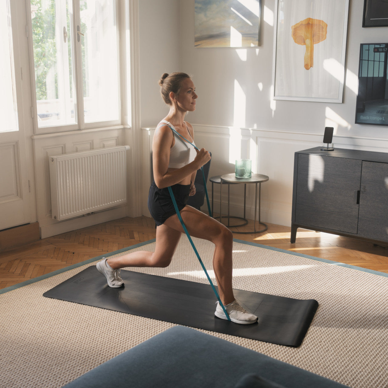 Frau macht Ausfallschritte mit Widerstandsbändern auf einer Yogamatte in einem hellen Wohnzimmer und nutzt dabei eine Functional-Fitness-Routine für ein effektives Ganzkörpertraining.