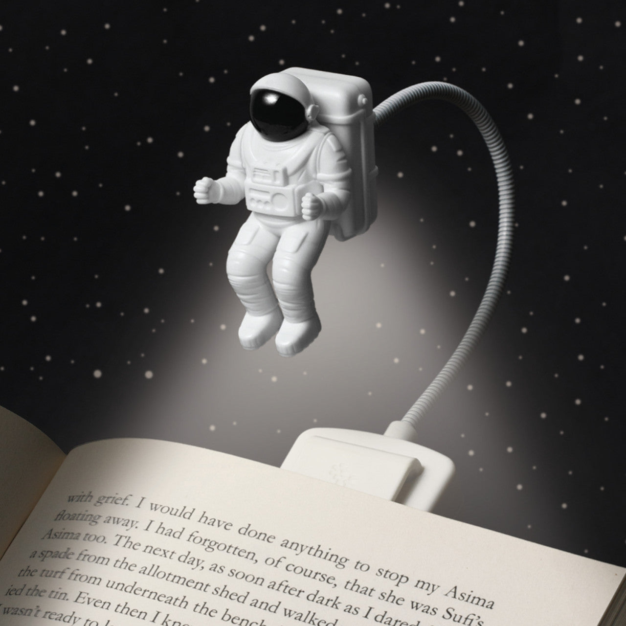 Eine kleine Astronauten-Leseleuchte schwebt über einem offenen Buch und ihr LED-Lichtstrahl beleuchtet die Seiten vor einem Sternenhintergrund.
