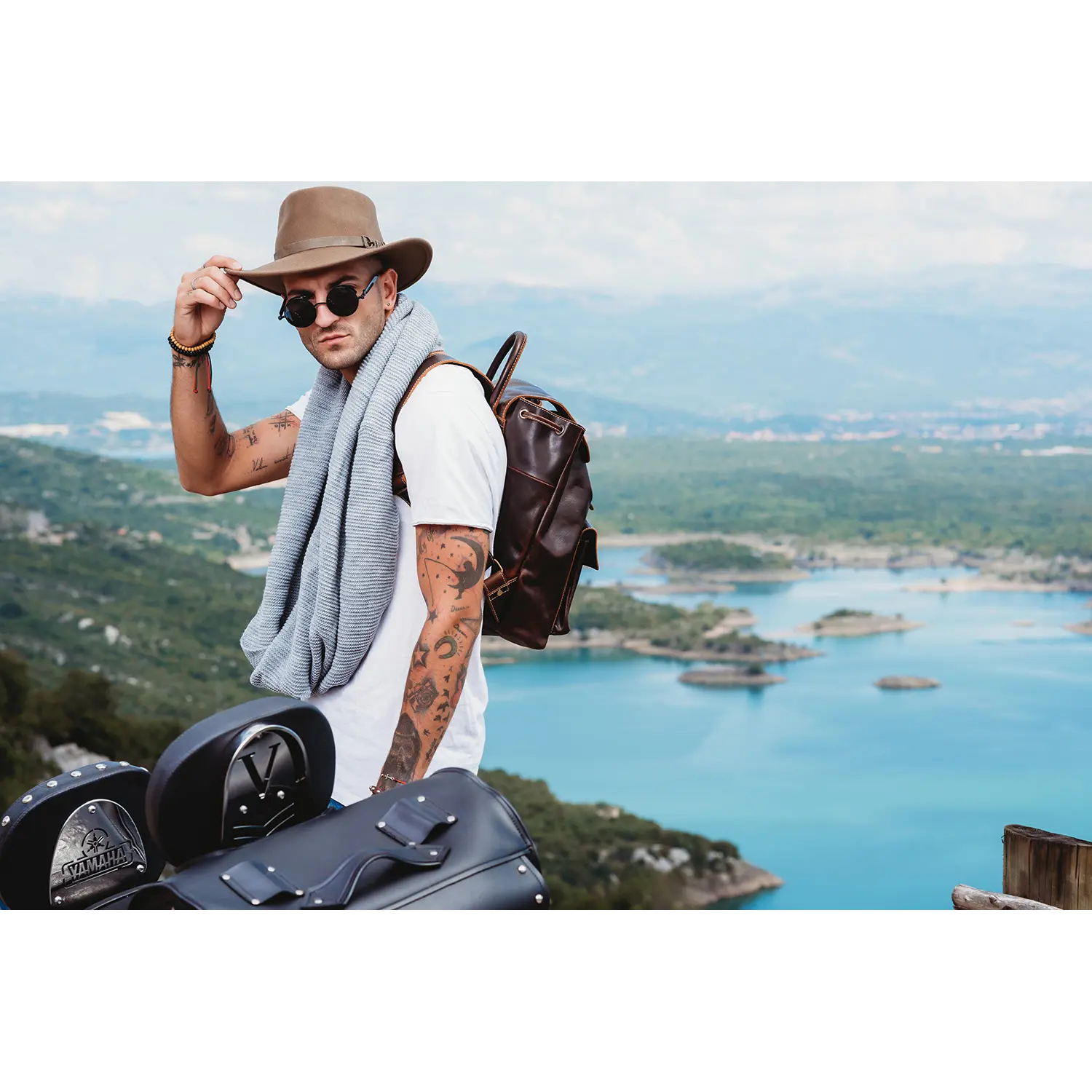 Ein Mann mit Hut und Sonnenbrille und einem Schal um den Hals hält die Krempe seines Hutes, während er neben Motorrädern steht und auf einen malerischen See und eine malerische Landschaft blickt. Er trägt den Vintage-Tagesrucksack JACOB'S WAY aus Leder.