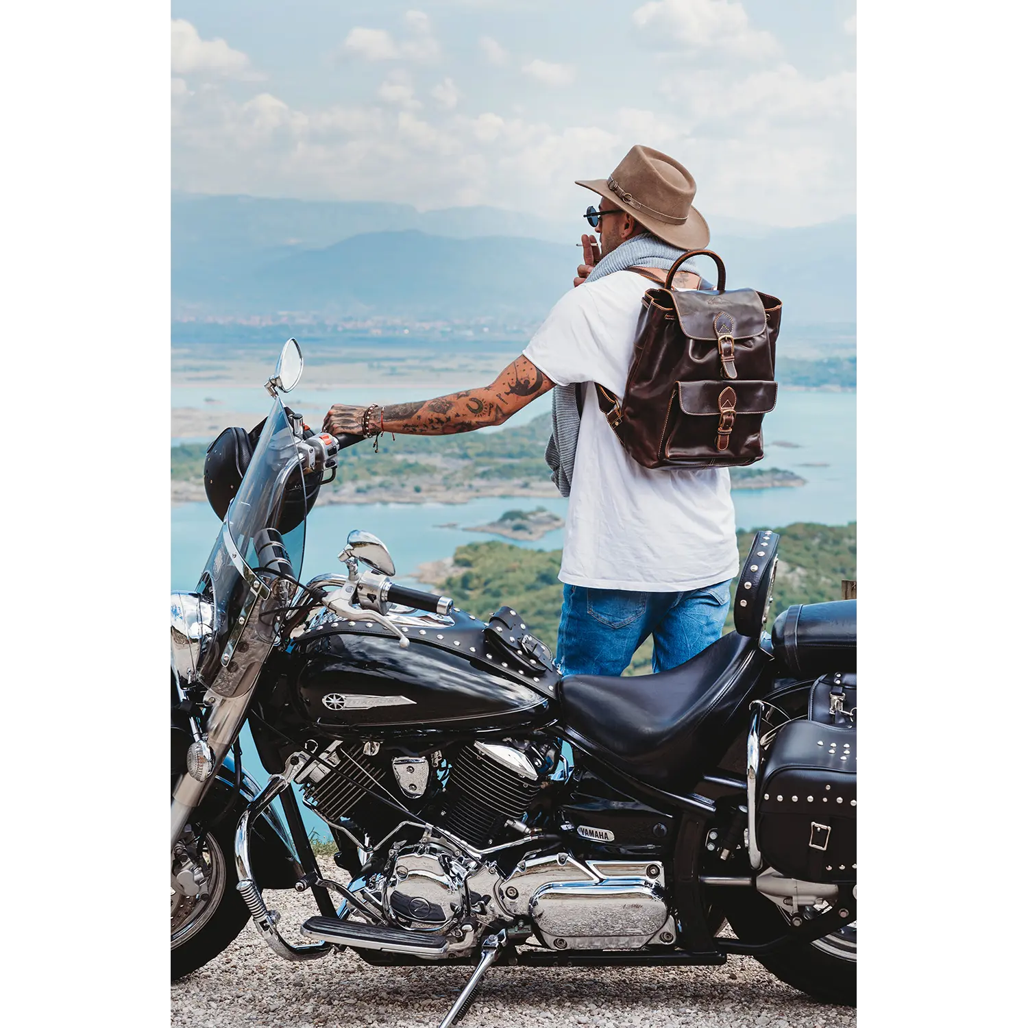 Ein Mann mit Hut und Jeans und Tattoos am Arm steht neben einem Motorrad und blickt auf den malerischen See und die Berge. Er hält eine Tasse in der Hand und trägt den Leder-Rucksack JACOB'S WAY. Sein weißes T-Shirt rundet das Bild unbeschwerter Abenteuerlust ab.