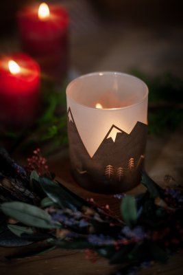 Brennende Kerze in einem Windlichthalter mit Bergmuster, mit roten Kerzen und verschwommenem Grün im Hintergrund.