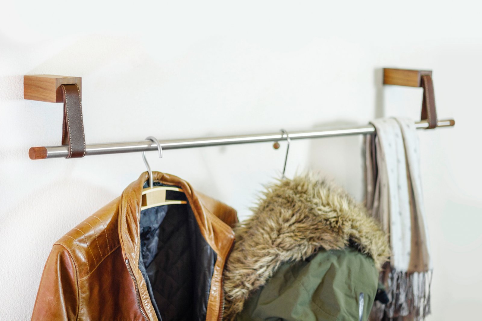 An einer Wandgarderobe aus Metall und Nussbaumholz hängen eine braune Jacke, ein grüner Parka und zwei Schals.