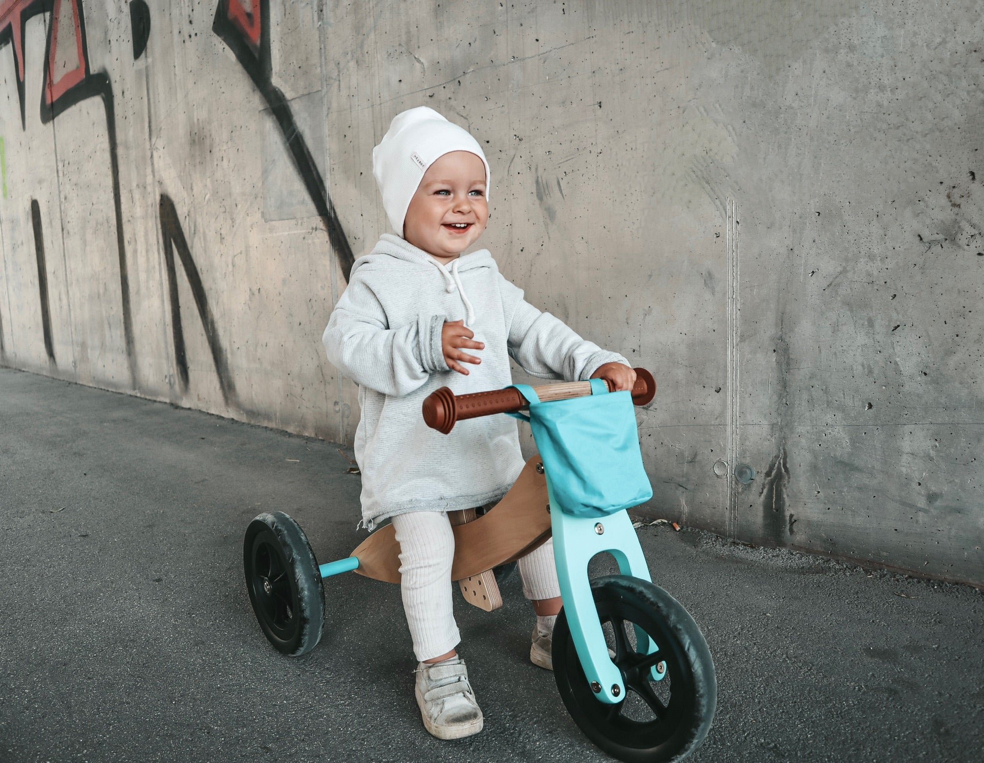 Ein lächelndes Kleinkind im grauen Outfit fährt auf einem blauen Lauflern-Trike an einer mit Graffiti bedeckten Wand entlang und verbessert mit jedem Anstoß seine motorischen Fähigkeiten.