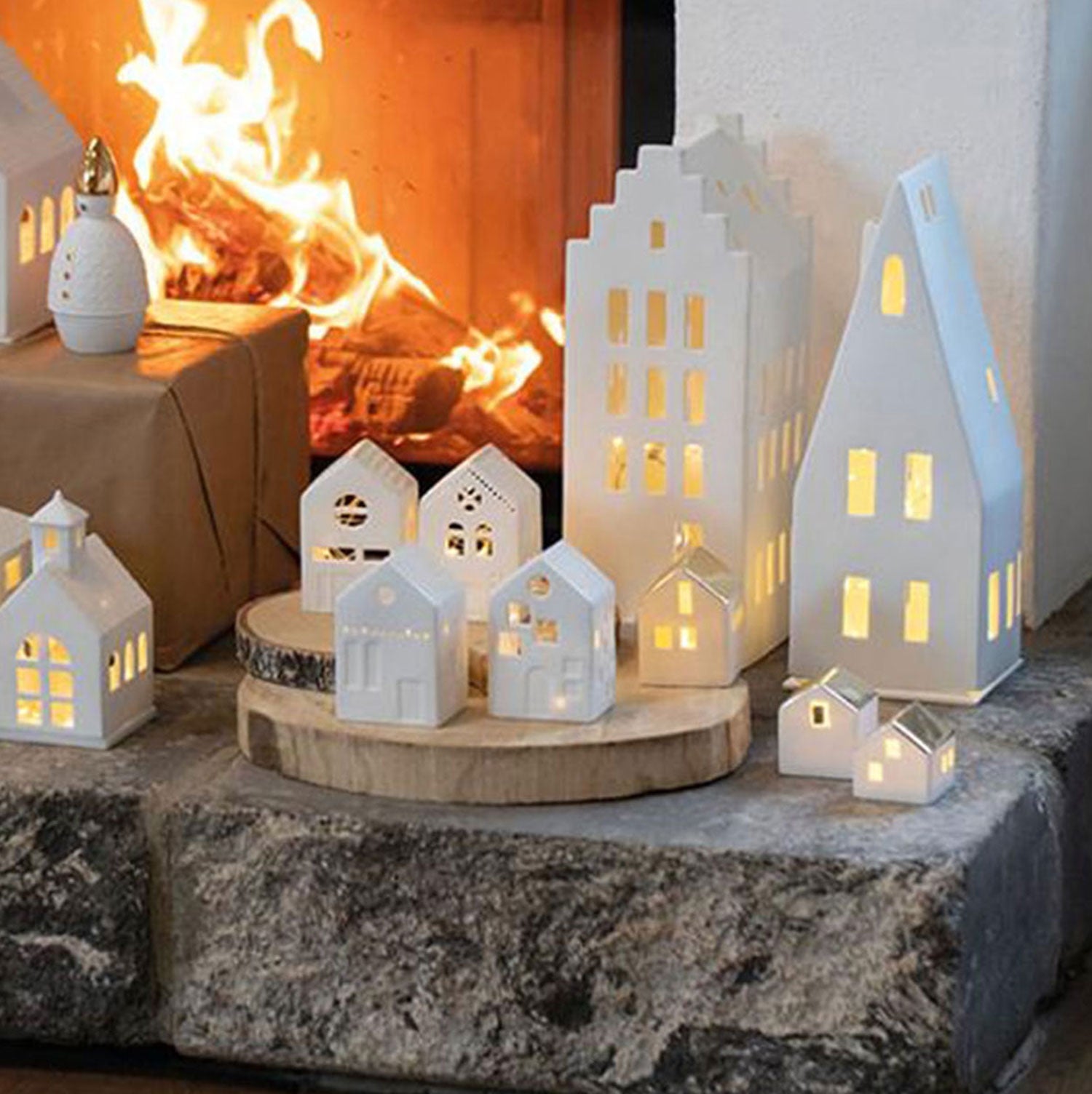 Das Große Lichthaus „Spitzdach“ aus unglasiertem Porzellan wird kunstvoll auf Holzscheiben am Kamin in Szene gesetzt. Das warme Feuerlicht verstärkt den gemütlichen, handgefertigten Charme dieser Szenerie.