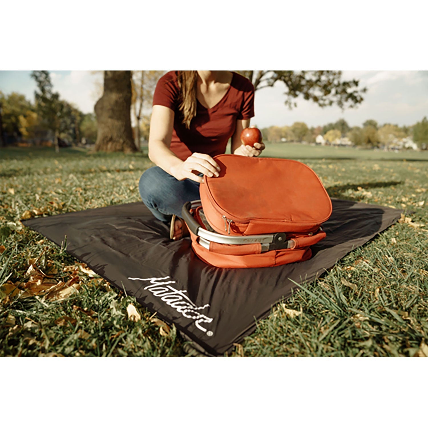 Person sitzt auf einer kompakten Outdoor-Decke im Park mit roter Tasche und Apfel.